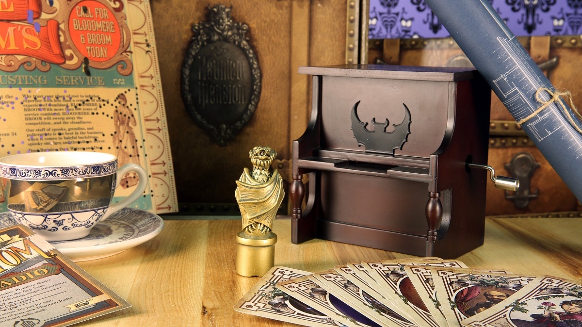 A styled flatlay of Ghost Post subscription box contents on a wooden surface: a miniature upright piano music box with bat-wing cutouts, a gold owl figurine, a spread of illustrated tarot-style playing cards, a teacup and saucer, and a vintage-style Ghost Post broadsheet, with a steamer trunk and rolled blueprints in the background.